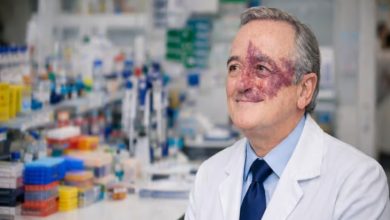 Laboratuvar ortamında, beyaz önlük ve kravatlı bir bilim insanı, arkasında deney malzemeleri bulunan tezgâh önünde otururken gülümseyerek kameranın dışına bakıyor. Yüzünün bir kısmında belirgin bir doğum lekesi görülüyor.