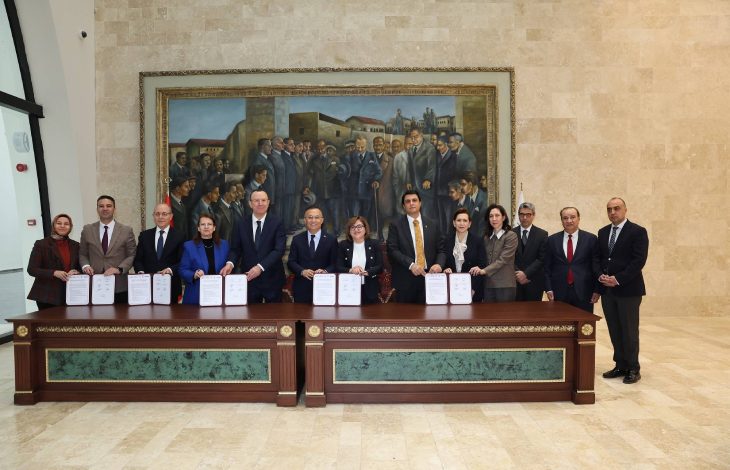 Protokol töreninde ayakta bulunan yetkililerin önünde uzun bir masa bulunmakta ve ellerinde tuttuklarını protokol dosyaları ile poz veriyorlar.