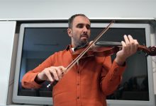 Bir sınıf ortamında erkek bir müzisyen ayakta keman çalıyor. Turuncu gömlekli kişi enstrümanı omzuna dayamış, yayı teller üzerinde dikkatle hareket ettiriyor. Arkasında büyük bir akıllı tahta bulunuyor; ortam ders ya da atölye havası taşıyor.