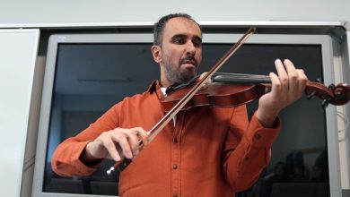 Bir sınıf ortamında erkek bir müzisyen ayakta keman çalıyor. Turuncu gömlekli kişi enstrümanı omzuna dayamış, yayı teller üzerinde dikkatle hareket ettiriyor. Arkasında büyük bir akıllı tahta bulunuyor; ortam ders ya da atölye havası taşıyor.