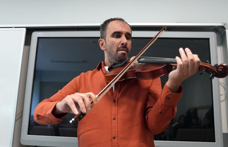 Bir sınıf ortamında erkek bir müzisyen ayakta keman çalıyor. Turuncu gömlekli kişi enstrümanı omzuna dayamış, yayı teller üzerinde dikkatle hareket ettiriyor. Arkasında büyük bir akıllı tahta bulunuyor; ortam ders ya da atölye havası taşıyor.