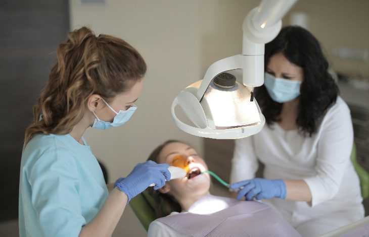 Bir diş kliniğinde hasta koltukta uzanmış şekilde tedavi görüyor. Maske ve eldiven takan iki sağlık çalışanı, güçlü bir muayene lambası altında hastanın ağzına müdahale ediyor. Ortam steril ve klinik bir atmosfer taşıyor.