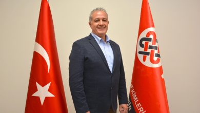 Görselde bir erkek resmi bir ortamda ayakta poz veriyor. Üzerinde lacivert bir blazer ceket ve açık renk gömlek var. Gülümseyerek kameraya bakıyor. Yanlarında iki bayrak bulunuyor: biri Türk bayrağı, diğeri ise kırmızı zemin üzerinde kurumsal bir logo ve yazı bulunan bir bayrak. Arka plan sade ve açık renkli bir duvardan oluşuyor.