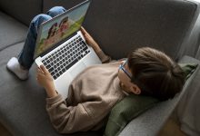 Görselde bir çocuk gri bir koltukta uzanmış halde dizüstü bilgisayar kullanıyor. Üzerinde açık kahverengi bir sweatshirt ve kot pantolon var, gözlük takıyor. Başının altında yeşil bir yastık bulunuyor. Bilgisayar ekranında çizgi film benzeri iki karakter görünüyor. Çocuk rahat bir pozisyonda ekrana odaklanmış görünüyor.