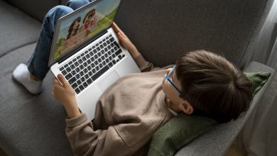 Görselde bir çocuk gri bir koltukta uzanmış halde dizüstü bilgisayar kullanıyor. Üzerinde açık kahverengi bir sweatshirt ve kot pantolon var, gözlük takıyor. Başının altında yeşil bir yastık bulunuyor. Bilgisayar ekranında çizgi film benzeri iki karakter görünüyor. Çocuk rahat bir pozisyonda ekrana odaklanmış görünüyor.