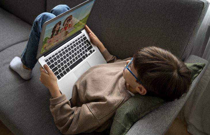 Görselde bir çocuk gri bir koltukta uzanmış halde dizüstü bilgisayar kullanıyor. Üzerinde açık kahverengi bir sweatshirt ve kot pantolon var, gözlük takıyor. Başının altında yeşil bir yastık bulunuyor. Bilgisayar ekranında çizgi film benzeri iki karakter görünüyor. Çocuk rahat bir pozisyonda ekrana odaklanmış görünüyor.