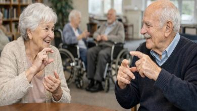 Bir huzurevinde, masada karşılıklı oturan iki yaşlı birey el hareketleriyle işaret diliyle sohbet ediyor; arka planda tekerlekli sandalyedeki diğer yaşlılar görülüyor.