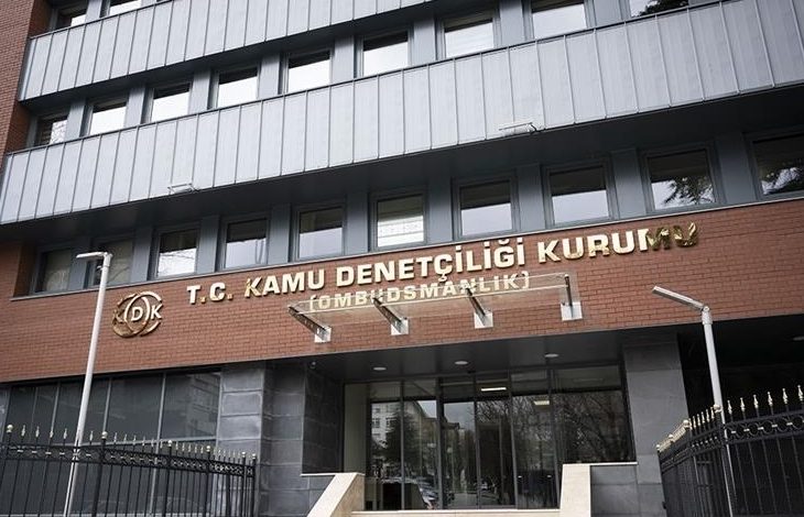 Çok katlı bir kamu binasının giriş cephesi görülüyor. Kapının üzerinde “T.C. Kamu Denetçiliği Kurumu (Ombudsmanlık)” yazısı yer alıyor. Binanın önünde demir parmaklıklar ve giriş merdivenleri bulunuyor. Resmi bir kurum binası izlenimi veren, dış mekânda çekilmiş bir fotoğraf.