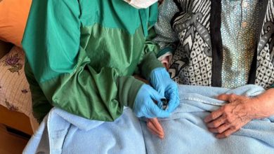 Bir ev ortamında sağlık çalışanı olduğu anlaşılan kişi, yeşil koruyucu önlük, maske ve eldivenle yaşlı bir kadının eline bakım uyguluyor. Yaşlı kadının üzeri battaniyeyle örtülü; işlem dikkatli ve özenli bir şekilde yapılıyor. Görsel, evde sağlık ve bakım hizmetini yansıtıyor.