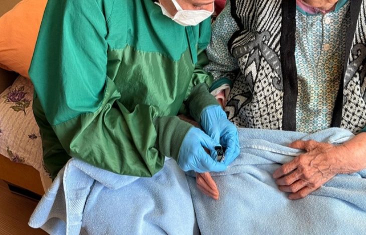 Bir ev ortamında sağlık çalışanı olduğu anlaşılan kişi, yeşil koruyucu önlük, maske ve eldivenle yaşlı bir kadının eline bakım uyguluyor. Yaşlı kadının üzeri battaniyeyle örtülü; işlem dikkatli ve özenli bir şekilde yapılıyor. Görsel, evde sağlık ve bakım hizmetini yansıtıyor.