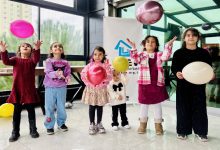 Camla çevrili bir alanda, farklı yaşlarda altı çocuk renkli balonlarla oynuyor. Çocuklar gülümseyerek balonları havaya atarken arkada bir dernek logosu bulunan afiş yer alıyor; ortam neşeli ve kutlama havasında.