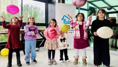 Camla çevrili bir alanda, farklı yaşlarda altı çocuk renkli balonlarla oynuyor. Çocuklar gülümseyerek balonları havaya atarken arkada bir dernek logosu bulunan afiş yer alıyor; ortam neşeli ve kutlama havasında.