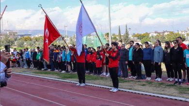Bir atletizm pistinde tören düzenlenmiş. Önde iki sporcu Türk bayrağı ve organizasyon bayrağını tutuyor. Arkalarında sporcular ve katılımcılar sıra halinde dizilmiş, saygı duruşu ya da açılış anı gibi resmi bir atmosfer var.