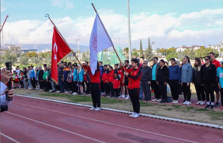 Bir atletizm pistinde tören düzenlenmiş. Önde iki sporcu Türk bayrağı ve organizasyon bayrağını tutuyor. Arkalarında sporcular ve katılımcılar sıra halinde dizilmiş, saygı duruşu ya da açılış anı gibi resmi bir atmosfer var.