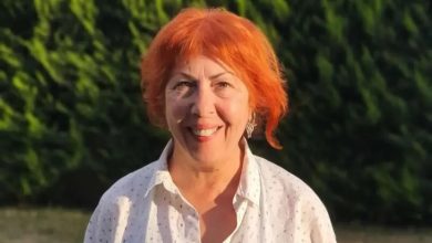 Açık havada, yeşil ağaçların önünde çekilmiş bir portre fotoğrafı. Kısa ve turuncu tonlarda saçlara sahip bir kadın, beyaz gömlekle kameraya doğru gülümseyerek poz veriyor. Gün ışığı yüzünü aydınlatıyor; arka plan doğal ve sakin bir ortam izlenimi veriyor.