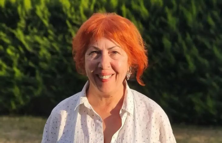 Açık havada, yeşil ağaçların önünde çekilmiş bir portre fotoğrafı. Kısa ve turuncu tonlarda saçlara sahip bir kadın, beyaz gömlekle kameraya doğru gülümseyerek poz veriyor. Gün ışığı yüzünü aydınlatıyor; arka plan doğal ve sakin bir ortam izlenimi veriyor.