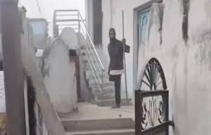 Dar ve taş bir geçitte, merdivenlerin bulunduğu eski bir yapı görülüyor. Merdiven başında elinde sopa benzeri bir nesne tutan bir kişi ayakta duruyor; çevrede duvarlar, metal korkuluklar ve giriş kapıları yer alıyor.