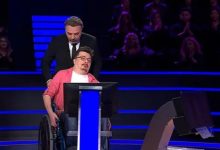 Bir televizyon yarışma programı stüdyosunda, tekerlekli sandalyede oturan bir yarışmacı ekran karşısında bulunuyor. Arkasında sunucu ayakta duruyor. Sahne mavi ışıklarla aydınlatılmış; arka planda seyirci sıraları seçiliyor. Yarışmacı duygusal bir an yaşıyor gibi görünürken, sunucu destekleyici bir duruş sergiliyor.