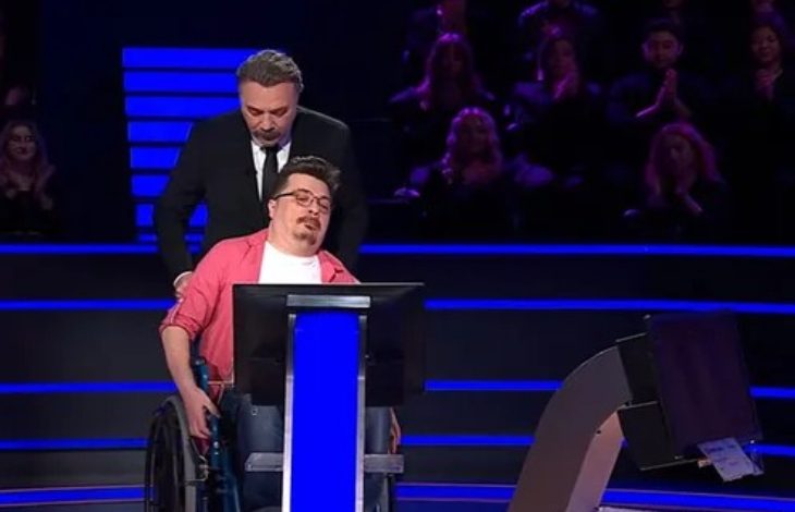 Bir televizyon yarışma programı stüdyosunda, tekerlekli sandalyede oturan bir yarışmacı ekran karşısında bulunuyor. Arkasında sunucu ayakta duruyor. Sahne mavi ışıklarla aydınlatılmış; arka planda seyirci sıraları seçiliyor. Yarışmacı duygusal bir an yaşıyor gibi görünürken, sunucu destekleyici bir duruş sergiliyor.