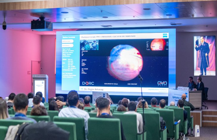 Görselde, bir konferans salonunda oturan izleyicilerin önünde büyük bir ekranda tıbbi bir sunum yapılırken, sahnede konuşmacıların retina görüntüleri içeren bir slaytı anlattığı görülüyor.