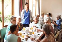 Aydınlık bir yemek salonunda, bir bakıcının eşliğinde masada birlikte kahvaltı yapan yaşlı bir grup insan görülüyor.
