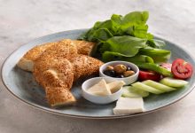 Kahvaltı tabağında sosyete simidi, karışık zeytin, domates, salatalık, üçgen krem peynir, beyaz peynir, roka yaprakları bulunuyor.