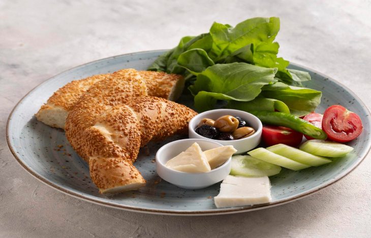 Kahvaltı tabağında sosyete simidi, karışık zeytin, domates, salatalık, üçgen krem peynir, beyaz peynir, roka yaprakları bulunuyor.