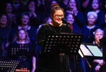 Sahnede mikrofon başında duran bir solist, notalık önünde gülümseyerek şarkı söylüyor. Arka planda koro üyeleri yer alıyor; ışıklandırma ve kalabalık, coşkulu bir konser atmosferi yaratıyor.