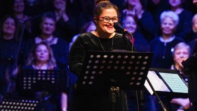 Sahnede mikrofon başında duran bir solist, notalık önünde gülümseyerek şarkı söylüyor. Arka planda koro üyeleri yer alıyor; ışıklandırma ve kalabalık, coşkulu bir konser atmosferi yaratıyor.