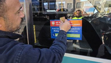 Bir kişi, şehir içi bir otobüsün camına bilgilendirici bir afiş yapıştırıyor. Camda yansıyan belediye görevlisi ve arka plandaki caddeyle birlikte sahne, toplu taşımada farkındalık ve denetim çalışmasını çağrıştırıyor.