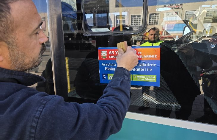 Bir kişi, şehir içi bir otobüsün camına bilgilendirici bir afiş yapıştırıyor. Camda yansıyan belediye görevlisi ve arka plandaki caddeyle birlikte sahne, toplu taşımada farkındalık ve denetim çalışmasını çağrıştırıyor.