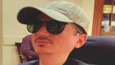 Şapka ve güneş gözlüğü takan bir erkek, kapalı bir mekânda yandan profilden otururken görülüyor.