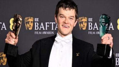 Siyah smokin ve papyon giymiş bir erkek, arkasında “EE BAFTA Film Awards” yazılı fon önünde poz veriyor. Her iki elinde de BAFTA maskesi şeklindeki ödülleri kaldırmış, gülümseyerek kameraya bakıyor.