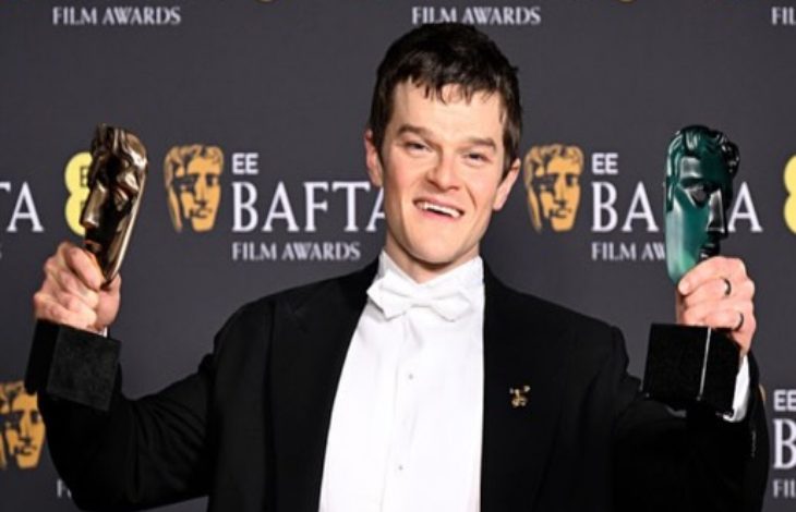 Siyah smokin ve papyon giymiş bir erkek, arkasında “EE BAFTA Film Awards” yazılı fon önünde poz veriyor. Her iki elinde de BAFTA maskesi şeklindeki ödülleri kaldırmış, gülümseyerek kameraya bakıyor.