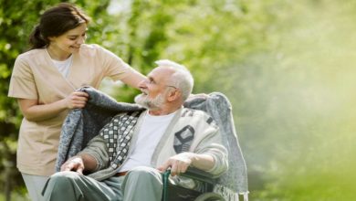 Açık havada, yeşillikler içinde bir park ortamında tekerlekli sandalyede oturan yaşlı bir adamın omuzlarına genç bir kadın bakım görevlisi tarafından battaniye örtülüyor. İkisi de gülümsüyor; sahne sıcak, şefkatli ve huzurlu bir bakım anını yansıtıyor.