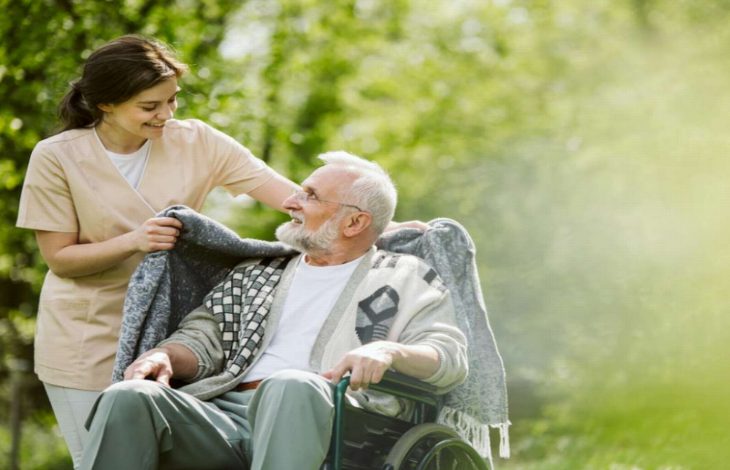Açık havada, yeşillikler içinde bir park ortamında tekerlekli sandalyede oturan yaşlı bir adamın omuzlarına genç bir kadın bakım görevlisi tarafından battaniye örtülüyor. İkisi de gülümsüyor; sahne sıcak, şefkatli ve huzurlu bir bakım anını yansıtıyor.