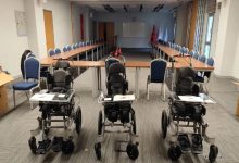 Bir toplantı salonunda U düzeninde yerleştirilmiş uzun masalar ve sandalyeler görülüyor. Salonun ön kısmında yan yana dizilmiş üç adet çocuklara yönelik özel tekerlekli sandalye/pozisyonlama cihazı bulunuyor. Arka tarafta projeksiyon perdesi ve Türk bayrakları yer alıyor. Ortam resmi bir tanıtım ya da teslim töreni havası veriyor.