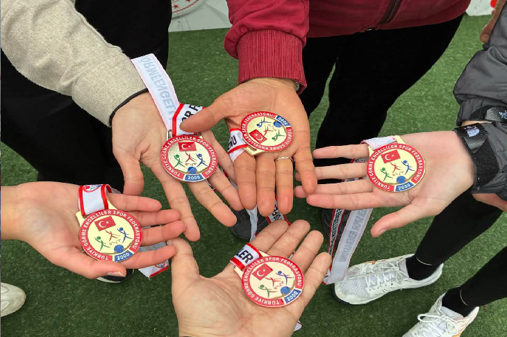 Spor sahasında beş kişi avuçlarını açmış, ellerinde madalyalar var. Madalyaların üzerinde Türk bayrağı ve federasyon logosu görülüyor. Arkada çim zemin ve spor kıyafetleri dikkat çekiyor; ödül anının sıcak bir hatırası gibi.