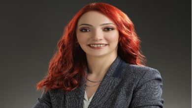 Görselde, profesyonel bir stüdyo ortamında çekilmiş bir portre fotoğrafı bulunuyor. Kırmızı saçlı, güler yüzlü bir kadın kamera karşısında poz veriyor. Üzerinde koyu renk bir blazer ceket ve açık renk bir bluz var. Kollarını hafifçe çaprazlayarak kendinden emin ve samimi bir duruş sergiliyor. Arka plan sade ve koyu tonlarda, bu da kişinin ön planda görünmesini sağlıyor.