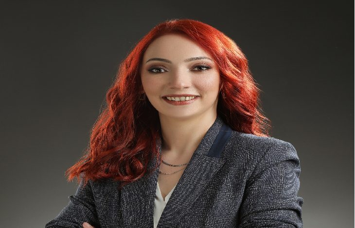 Görselde, profesyonel bir stüdyo ortamında çekilmiş bir portre fotoğrafı bulunuyor. Kırmızı saçlı, güler yüzlü bir kadın kamera karşısında poz veriyor. Üzerinde koyu renk bir blazer ceket ve açık renk bir bluz var. Kollarını hafifçe çaprazlayarak kendinden emin ve samimi bir duruş sergiliyor. Arka plan sade ve koyu tonlarda, bu da kişinin ön planda görünmesini sağlıyor.