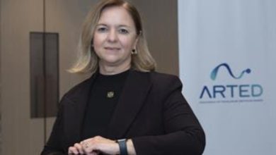 Görselde, kurumsal bir ortamda çekilmiş bir portre yer alıyor. Orta yaşlı bir kadın, siyah blazer ceket ve siyah üstle, bir masa ya da banko önünde poz vermiş. Arka planda sade bir ofis duvarı ve üzerinde “ARTED” yazılı bir logo bulunan bir pano görülüyor. Kadın kameraya doğrudan bakıyor ve hafif bir gülümsemeyle resmi bir duruş sergiliyor.