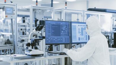 Görselde modern ve steril bir laboratuvar ortamı görülüyor. Beyaz koruyucu tulum, maske ve eldiven giymiş bir kişi bilgisayar başında çalışıyor. Önünde iki geniş monitör bulunuyor; ekranlarda teknik veriler ve analiz arayüzleri açık. Yan tarafta bir mikroskop yer alıyor. Arka planda cam bölmelerle ayrılmış, yüksek teknoloji ekipmanlarla donatılmış bir araştırma alanı dikkat çekiyor.