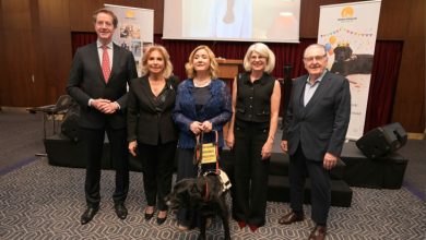 Bir konferans salonunda beş yetişkin yan yana poz veriyor. Ortadaki kadının yanında tasmalı siyah bir rehber köpek bulunuyor. Arkada sahne, projeksiyon ekranı ve etkinlik afişleri yer alıyor. Resmî ve anlamlı bir tören havası dikkat çekiyor.