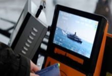 Bir toplu taşıma aracında yer alan elektronik kart okuma cihazı görülüyor. Ekranda deniz üzerinde ilerleyen bir vapur görüntüsü yer alıyor. Ön planda bir kişi ulaşım kartını cihaza tutuyor. Görsel, toplu taşımada kartlı geçiş sistemini ve elektronik bilet uygulamasını yansıtıyor.