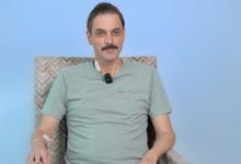 Açık mavi arka plan önünde bir koltukta oturan, oyuncu Ufuk Özkan açık yeşil tişört giymiş ve yakasında küçük bir mikrofon takılı şekilde, kolunda ve boynunda damar yolu pansumanları bulunarak kameraya doğru bakıyor.