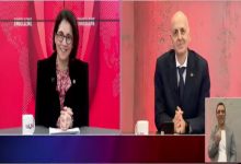 Bölünmüş ekranlı bir televizyon yayınında iki sunucu stüdyoda karşılıklı yer alıyor. Sol tarafta kadın sunucu masa başında gülümserken, sağda erkek konuk kameraya dönük oturuyor. Ekranın köşesinde işaret dili tercümanı bulunuyor. Kırmızı tonların hâkim olduğu bir haber programı atmosferi var.