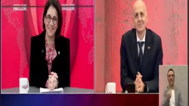 Bölünmüş ekranlı bir televizyon yayınında iki sunucu stüdyoda karşılıklı yer alıyor. Sol tarafta kadın sunucu masa başında gülümserken, sağda erkek konuk kameraya dönük oturuyor. Ekranın köşesinde işaret dili tercümanı bulunuyor. Kırmızı tonların hâkim olduğu bir haber programı atmosferi var.