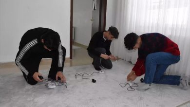 Bir odada üç genç yerde diz çökmüş şekilde elektrik kabloları ve küçük cihazlarla uğraşıyor. Önlerinde uzatma kablosu ve bağlantı aparatları bulunuyor. Oda sade; beyaz duvarlar ve tül perde arka planda görülüyor. Gençler bir cihazın kurulumunu ya da bağlantısını yapıyor gibi görünüyor.