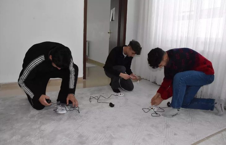 Bir odada üç genç yerde diz çökmüş şekilde elektrik kabloları ve küçük cihazlarla uğraşıyor. Önlerinde uzatma kablosu ve bağlantı aparatları bulunuyor. Oda sade; beyaz duvarlar ve tül perde arka planda görülüyor. Gençler bir cihazın kurulumunu ya da bağlantısını yapıyor gibi görünüyor.