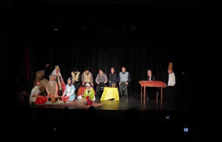 Bir tiyatro sahnesinde, siyah perde önünde geleneksel kıyafetler giymiş kalabalık bir oyuncu grubu yerde oturup sohbet ederken, sahnenin sağ tarafında masada oturan ve uzun külah şapka takan bir karakter dikkat çekiyor.